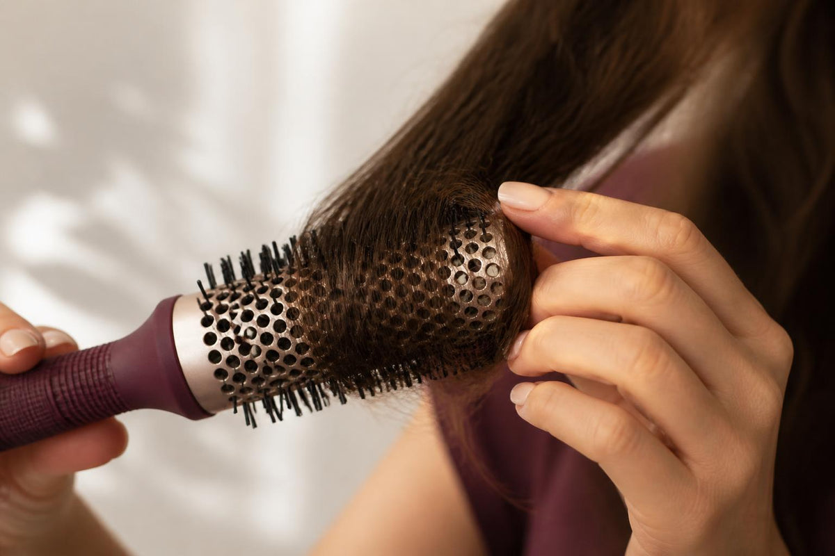 A woman with long, shiny hair applying a protective spray from a bottle before using a heated styling tool