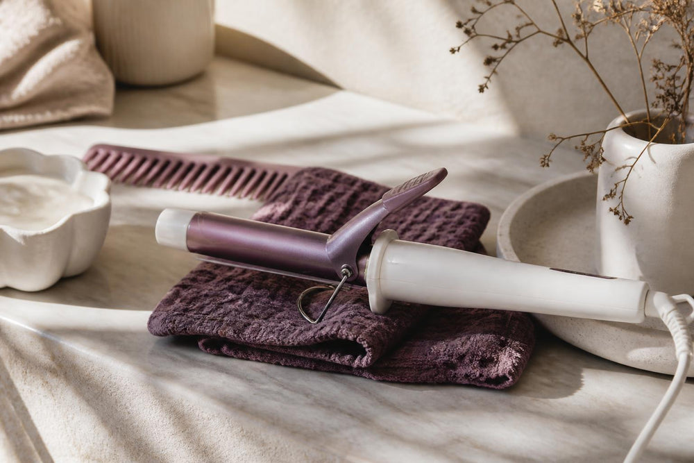 A collection of hair styling tools including a blow dryer, round brush, and curling iron arranged on a marble surface