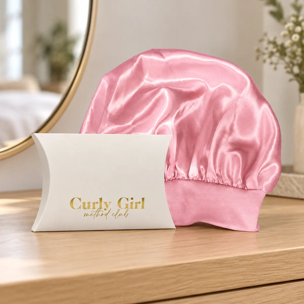 Pink satin sleep cap with its source packaging on a wooden dressing table.